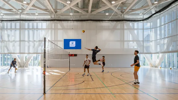Volleyball players mid-rally in a bright, glass-walled gymnasium